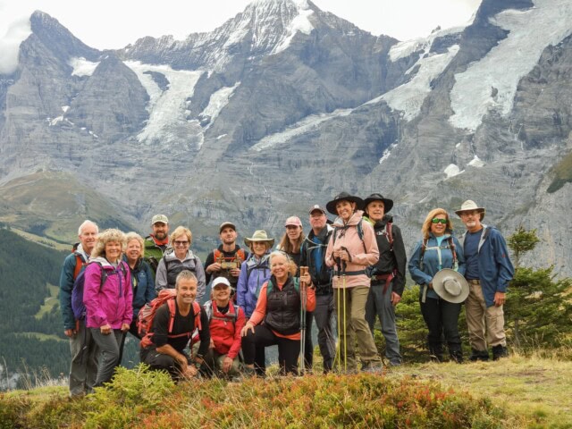 A group of 15 hikers, dressed in outdoor gear, pose for a photo in front of a mountainous landscape with snow-capped peaks and rocky terrain, capturing the essence of travel and tourism in Switzerland.