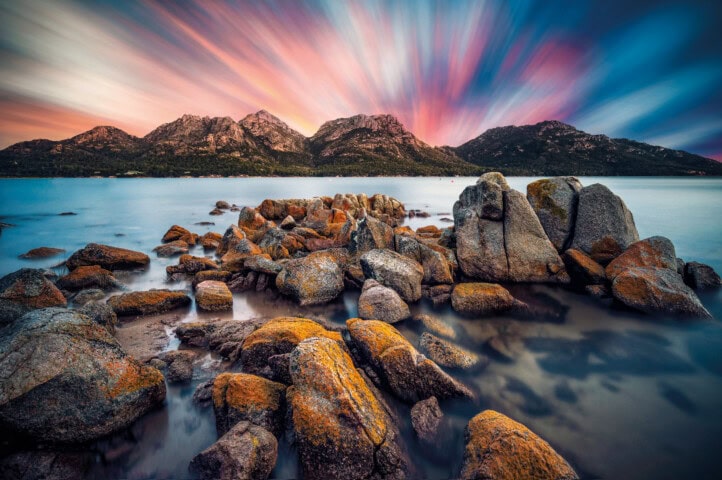 A serene Tasmanian landscape featuring a rocky shoreline with large boulders in the foreground, a calm body of water, and a backdrop of mountains under a vibrant, colorful sky.