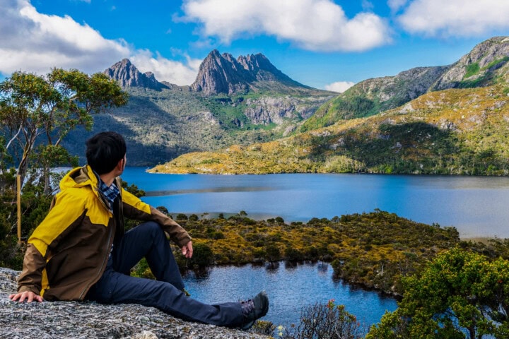 A person in a yellow jacket sits on a rock, overlooking a vibrant blue lake with mountainous terrain and a clear sky that calls to mind the pristine beauty of Tasmania.