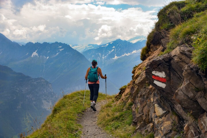 A hiker with trekking poles walks along a narrow mountain path in Switzerland, enjoying a scenic view of peaks under a partly cloudy sky; a red and white trail marker is painted on a rock, making it clear this route is popular for travel and tourism enthusiasts alike.