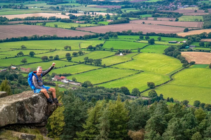 Male hiker smiling and waving while sitting on a rock perched over an English countryside valley in the North York Moors.