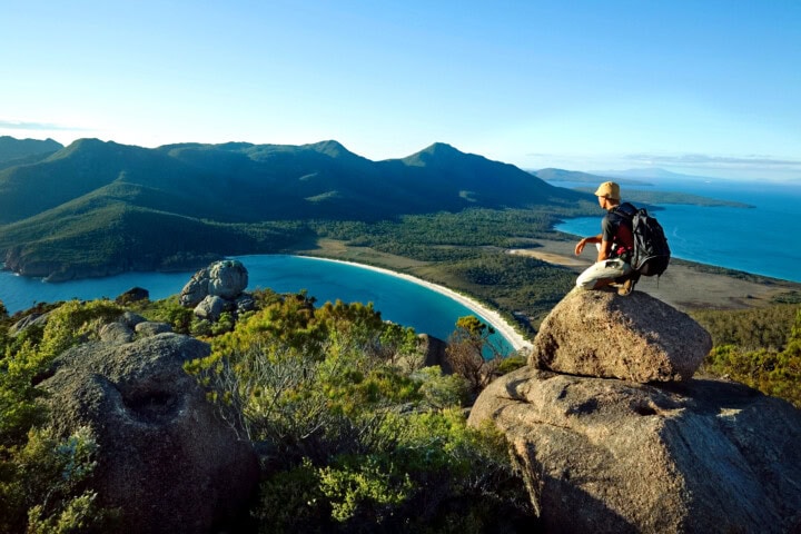 A hiker with a backpack sits on a large rock, overlooking a scenic view of Tasmania's beach, forest, and mountainous landscape.