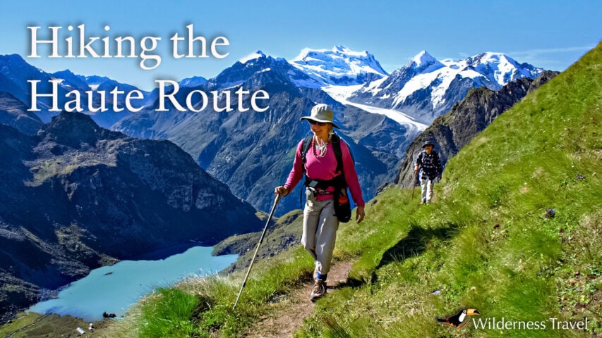 Two hikers walk along a narrow mountain trail with snow-capped peaks and a turquoise lake in the background, under clear blue skies. Text reads "Hiking the Haute Route".
