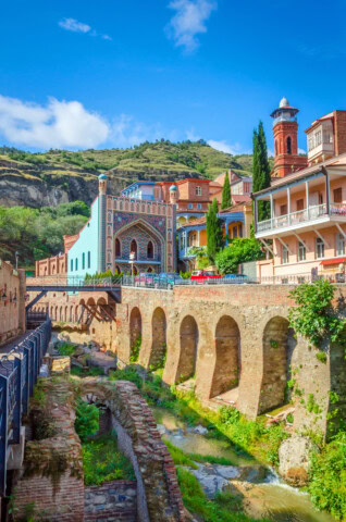 Colorful buildings and a mosque tower stand above a historical brick bridge over a small waterway in a scenic area of Georgia, surrounded by greenery and a hillside in the background—a perfect travel destination for those interested in tourism.