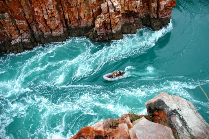 A small motorboat navigates through turbulent turquoise waters surrounded by high rocky cliffs, reminiscent of Australia's rugged coastline.