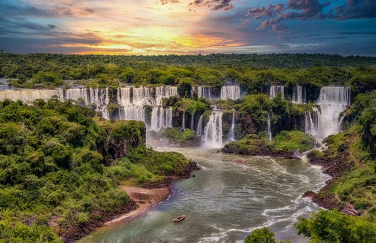 A vast landscape in Argentina shows multiple waterfalls pouring into a river surrounded by lush green trees under a vibrant sunset sky with scattered clouds.