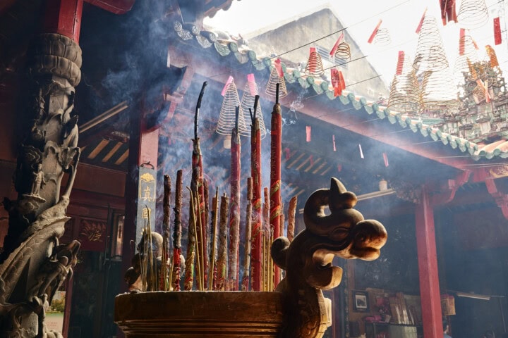 Numerous burning incense sticks with smoke rising in a large, decorative pot inside a traditional Vietnamese temple, adorned with hanging coils and red tags.