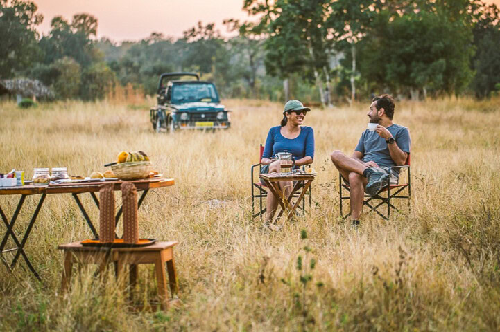 Two people, likely weary travelers, sit on chairs in a grassy field, engaged in a lively conversation. A parked vehicle is in the background, hinting at their recent hike or adventure. Tables laden with food and beverages are set up in the foreground, inviting them to relax and refresh.