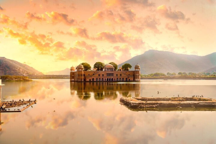 A grand palace with multiple domes stands in the middle of a calm lake surrounded by mountains at sunset, an ideal spot for an adventure tour, with birds perched on platforms in the foreground.