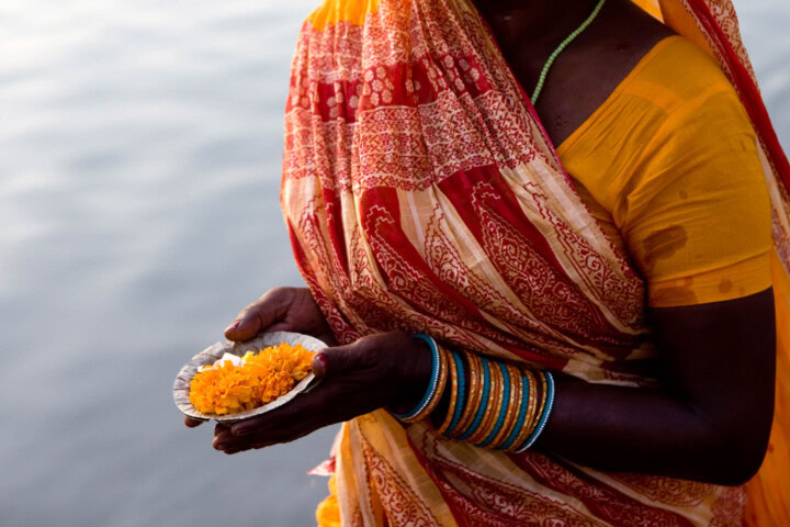 A person wearing a yellow and red sari holds a small dish with yellow flowers over water, capturing a serene moment on their adventure.