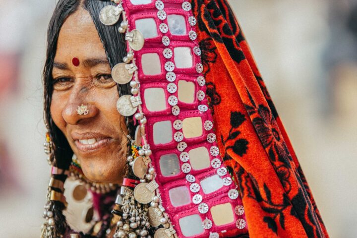 A woman wearing traditional, colorful attire with coin and mirror embellishments smiles, embodying the spirit of cultural adventure. She has a nose ring, a bindi on her forehead, and earrings as if preparing to be your vibrant guide on this unforgettable trip.