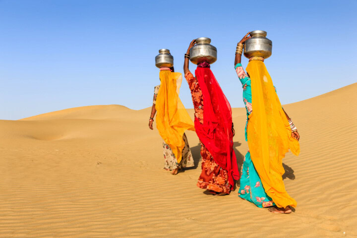 Three women in colorful sarees, like seasoned guides on a desert tour, carry metal pitchers on their heads while walking through a stunning landscape under a clear blue sky.