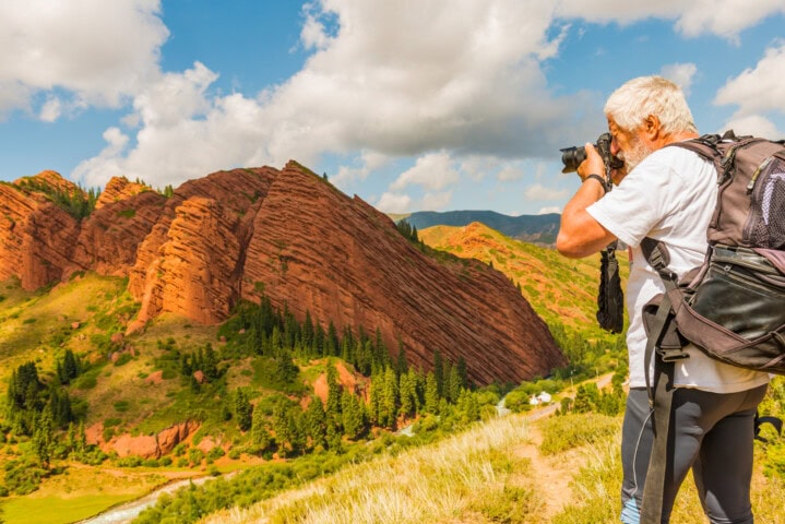 A person wearing a backpack and holding a camera is capturing the beauty of Kyrgyzstan's mountainous landscape, adorned with striking red rock formations on a sunny day.
