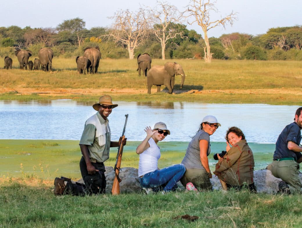 Group of five people sitting and posing for a photo near a lake with a herd of elephants in the background in a grassy, wooded area. The scenic beauty and diverse wildlife make it an ideal spot to visit all months of the year.