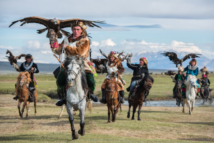 Horse riders in traditional Mongolian attire, each holding an eagle, ride across a grassy landscape with mountains in the background. They appear to be part of a cultural or hunting exhibition.