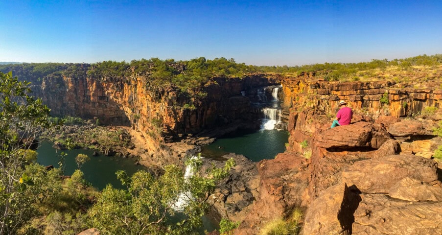 A person sits on a rocky outcrop overlooking a multi-tiered waterfall flowing into a river, surrounded by rugged cliffs and sparse vegetation under a clear blue sky—a perfect snapshot for those inspired by Australian tourism.