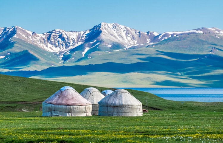 Three yurts are set up on a grassy plain near a lake with snow-capped mountains in the background under a clear blue sky, offering travelers an authentic Kyrgyzstan experience.