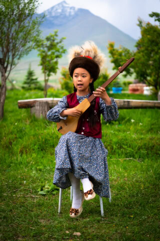 A young girl in traditional clothing plays a stringed instrument while seated outdoors, with the trees and snow-capped mountains of Kyrgyzstan in the background.