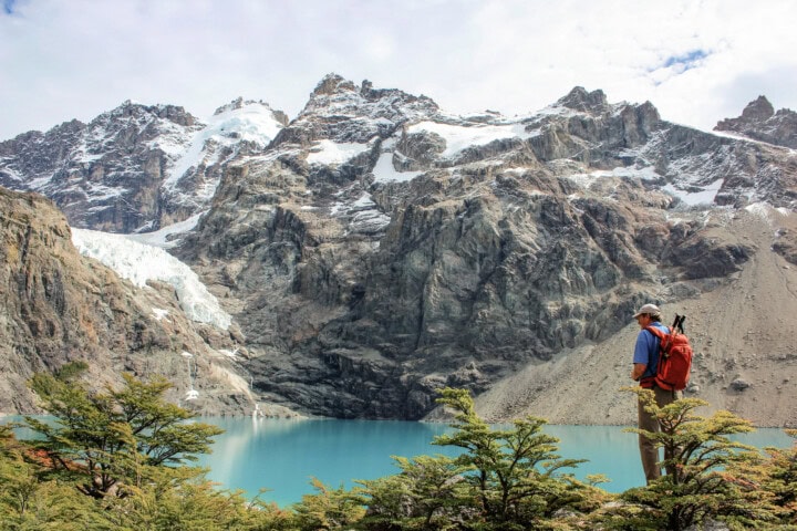 A person wearing a red backpack stands on the edge of a mountain lake in Argentina, with snow-capped peaks and rocky cliffs in the background.