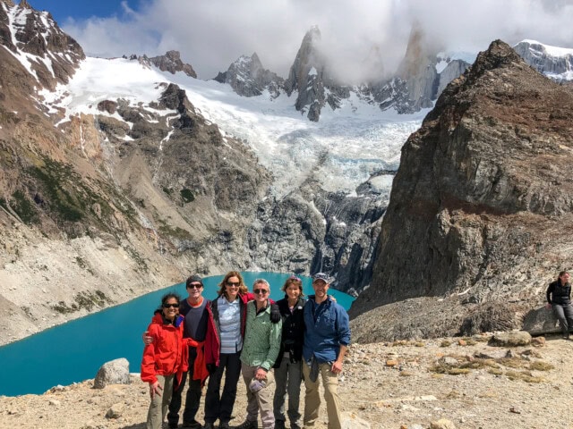 A group of six people stands together on rocky terrain in Argentina, with snow-capped mountains and a glacial lake in the background. Another person stands in the distance.