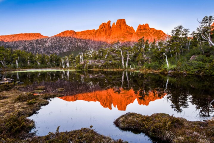 Mountain peaks in Tasmania illuminated by the setting sun, reflecting in a calm lake surrounded by trees and nature.