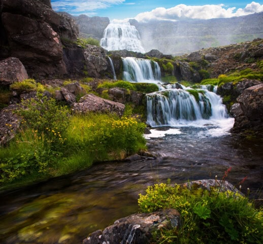 A cascading waterfall flows over rocks surrounded by green vegetation under a cloudy sky, making it a popular destination for travel and tourism in Iceland.