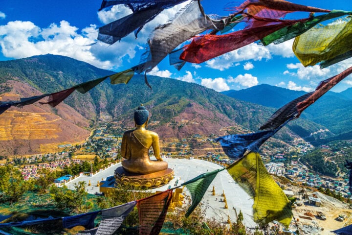 A large Buddha statue overlooks a valley town and mountains, with colorful prayer flags in the foreground fluttering in the wind.