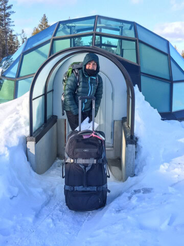 A person stands in front of an igloo-shaped glass building in Finland, dressed in winter attire and holding onto the handle of a large black suitcase. Snow surrounds the structure.