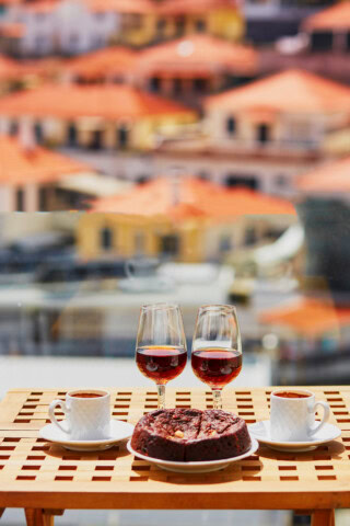 Two cups of coffee, two glasses of red wine, and a chocolate cake on a table, with blurred red-roofed buildings in the background hinting at a charming day in Portugal.