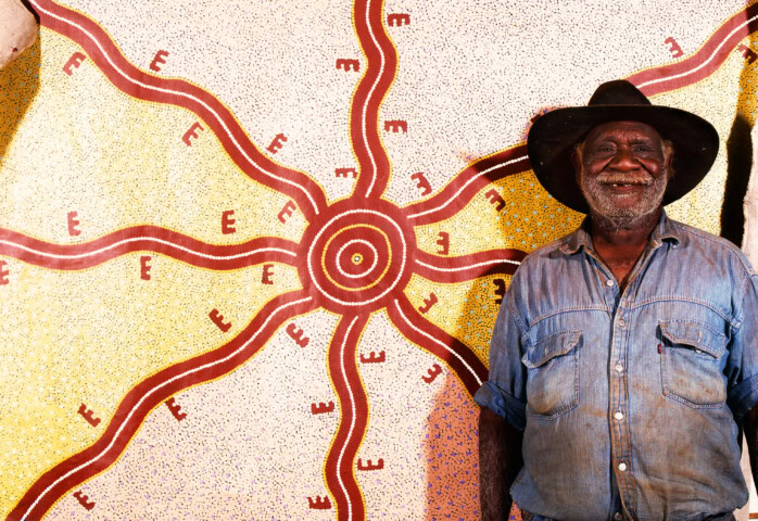 A man wearing a black hat and denim shirt stands smiling in front of an intricate and colorful Australian artwork featuring concentric circles and radiating lines.