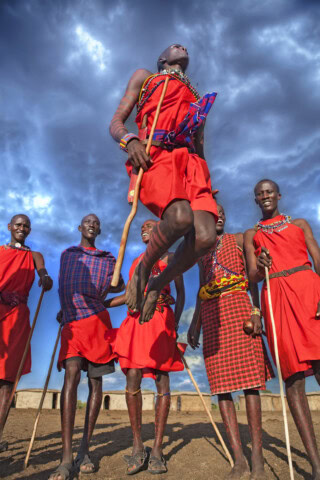 A group of individuals in traditional attire, with one person mid-air in a jump, captures the essence of their adventure. A cloudy sky looms in the background, adding to the thrill of their trip.
