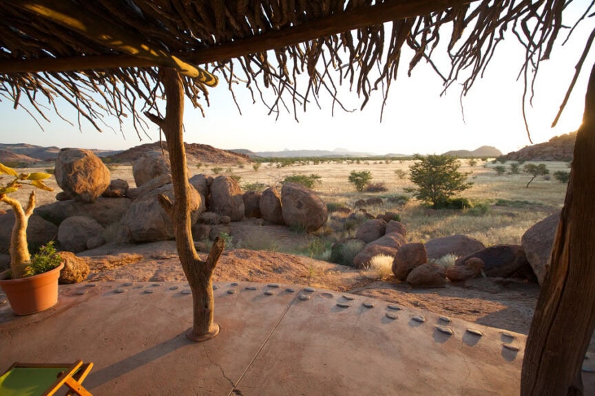 View from a shaded verandah overlooking a rocky landscape with scattered trees and grassy plains under a clear sky, evoking the adventurous spirit of Namibia's wonderland.