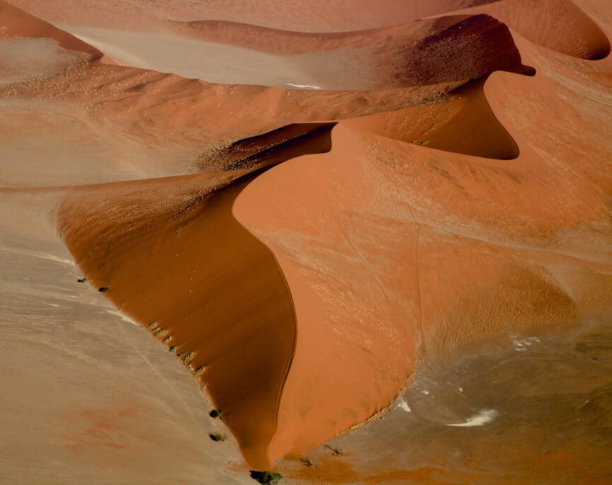 Aerial view of large, reddish sand dunes with smooth curves and ridges in a desert landscape, capturing the essence of Namibia's wonderland. Sparse vegetation is visible at the base of one dune.