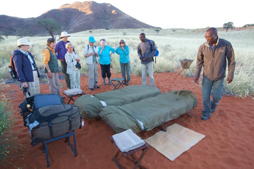 A group of adventurers in outdoor clothing stand around two sleeping bags set up on makeshift cots in a grassy, red sand area with the mountains of Namibia in the background. Camping gear is placed near them, adding to the sense of wonderland adventure.