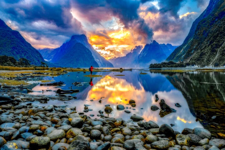 A serene lake in New Zealand with rocks in the foreground reflects mountainous scenery and a vibrant, colorful sky at sunrise or sunset, with a person standing on the shore in the distance.