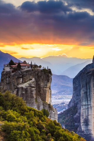 A monastery in Greece sits atop a steep cliff at sunset, with a dramatic sky and mountain range in the background. Trees cover the hillside leading up to the cliff.
