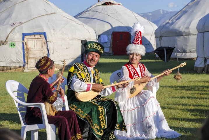 Three people in traditional attire play string instruments outdoors, with yurts and the majestic mountains of Mongolia in the background.