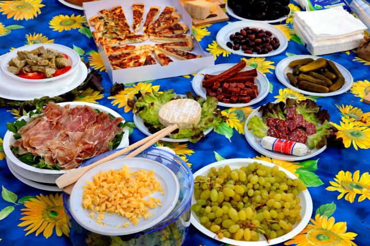A variety of foods including sliced meats, cheese, grapes, pickles, black olives, and pizza on a table with a blue sunflower-patterned tablecloth evokes the taste of travel in Switzerland.