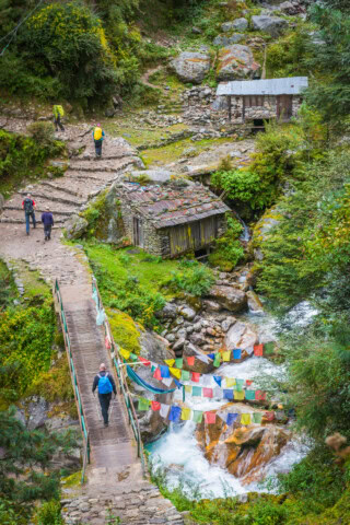 A couple of people crossing a bridge decorated with colorful prayer flags over a stream in Nepal. The area is surrounded by greenery, small houses, and stone pathways with a person in a yellow jacket ahead.