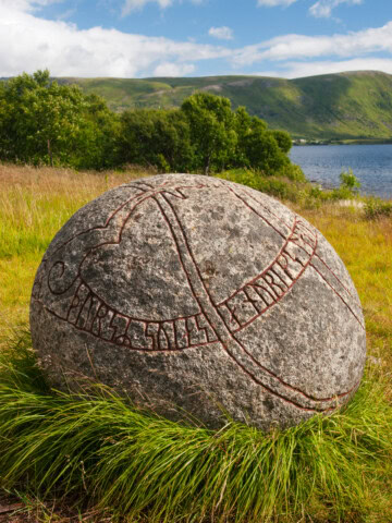 A large stone with engraved runes sits in a grassy field near a body of water, with green hills in the background under a partly cloudy sky, reminiscent of the serene landscapes found in Norway.