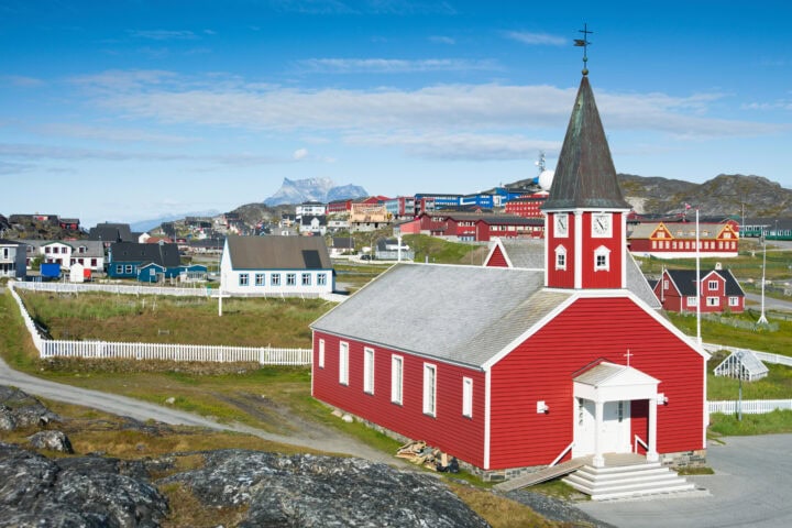 A red wooden church with a clock tower stands in the foreground, surrounded by colorful houses and rocky terrain under a blue sky.