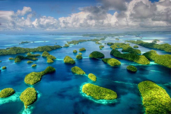 Aerial view of numerous lush, green islands surrounded by blue ocean under a partly cloudy sky, showcasing the paradisiacal allure of Palau for tourism and travel enthusiasts.