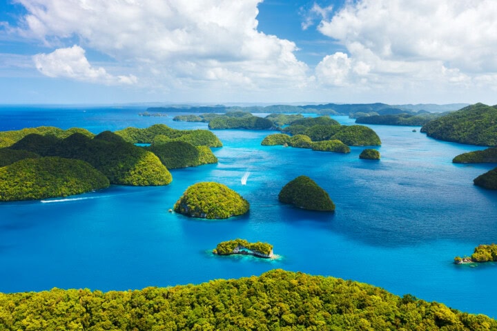 Aerial view of numerous green islands surrounded by clear blue ocean water under a partly cloudy sky, capturing the breathtaking beauty of Palau—a must-visit destination for travel and tourism enthusiasts.
