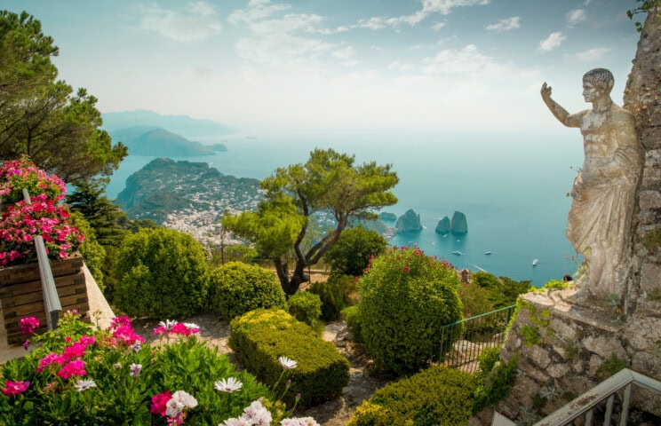 A scenic view of Italy's coastal landscape with lush greenery, vibrant flowers, a stone statue, and rocky islands in the blue sea under a clear sky.