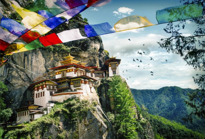 A monastery built on a rocky cliff in Bhutan is surrounded by lush mountains under a blue sky, with colorful prayer flags and flying birds in the foreground—a perfect scene for tourism and travel enthusiasts.