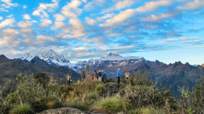 Hikers stand on a grassy hilltop with snow-capped mountains and a partly cloudy sky in the background, reminiscent of the breathtaking views seen when hiking the Inca Trail.