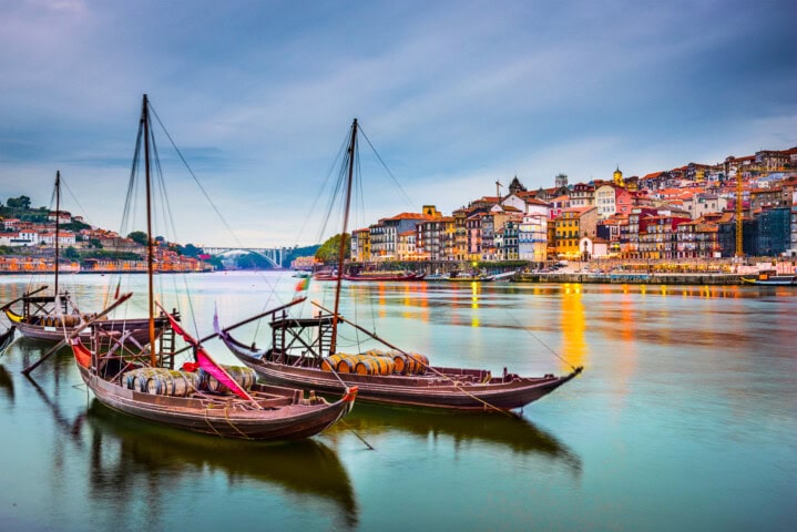 Boats gently float on the Douro River in Porto, Portugal, with colorful historic buildings illuminated by evening lights in the background.