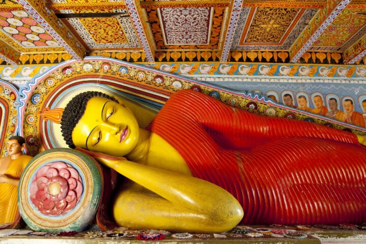A large reclining Buddha statue with a red robe, resting its head on a decorated cushion. The backdrop features vibrant, ornate patterns and smaller Buddha figures, reminiscent of the rich cultural heritage found in Sri Lanka.