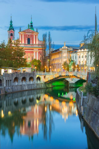 A serene view of Ljubljana's cityscape in Slovenia featuring the Ljubljanica River, the iconic pink Franciscan Church, and illuminated bridges and buildings at twilight.