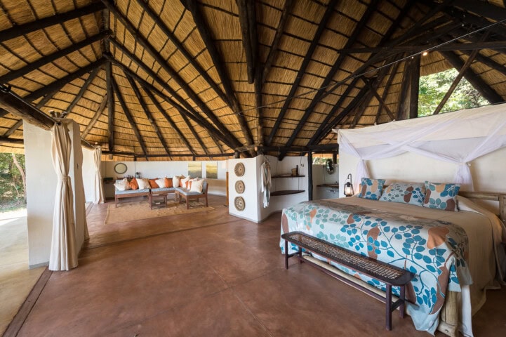 Spacious room with a thatched roof featuring a bed with a floral quilt, a rustic bench, a seating area with white cushions, and various decorations on the walls. The floor is made of polished brown concrete.